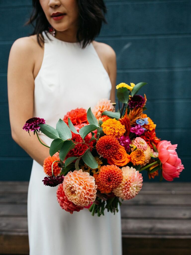 Bride holds a modern bouquet of orange, red, and blush pink dahlias. 