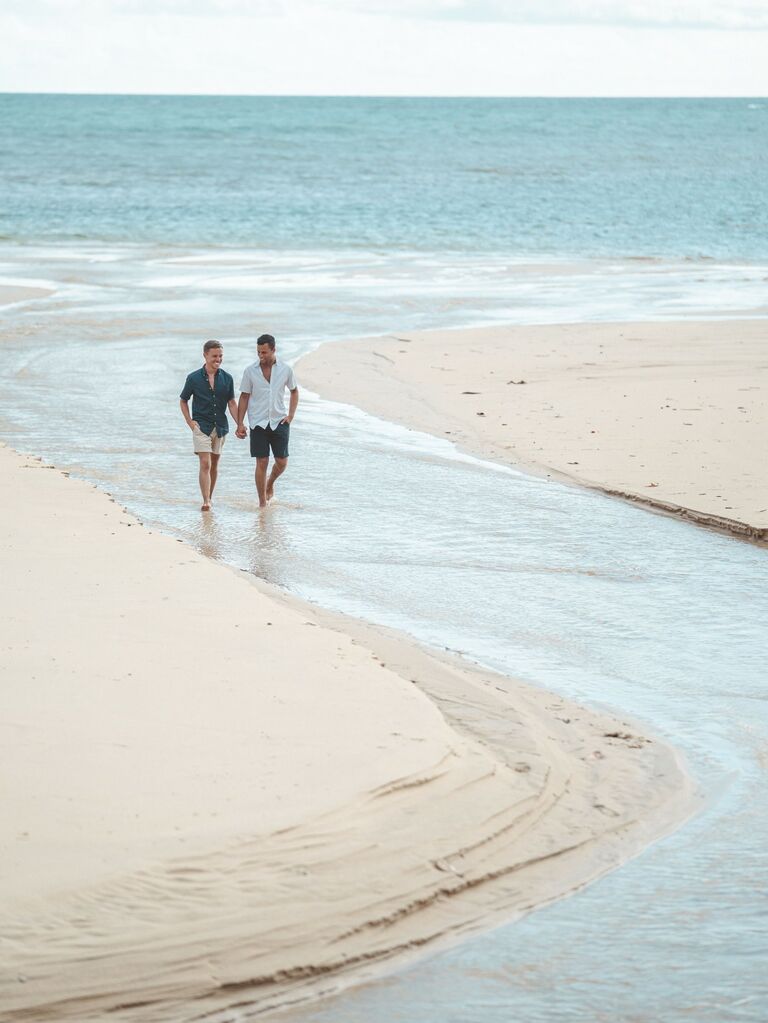 Couple walks through the water on a beach while holding hands. 