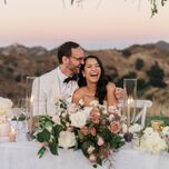 bride and groom laugh while listening to toast seated at outdoor sweetheart table decorated with light pink rose centerpiece, taper candles and mountain scenery in the background 
