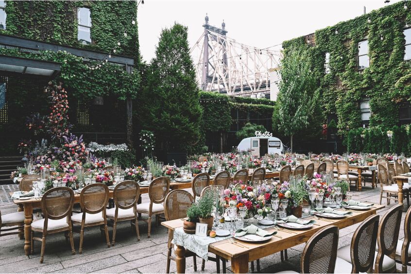 An outdoor rooftop, covered with greenery, features long tables set with florals and views of an industrial bridge in the background.