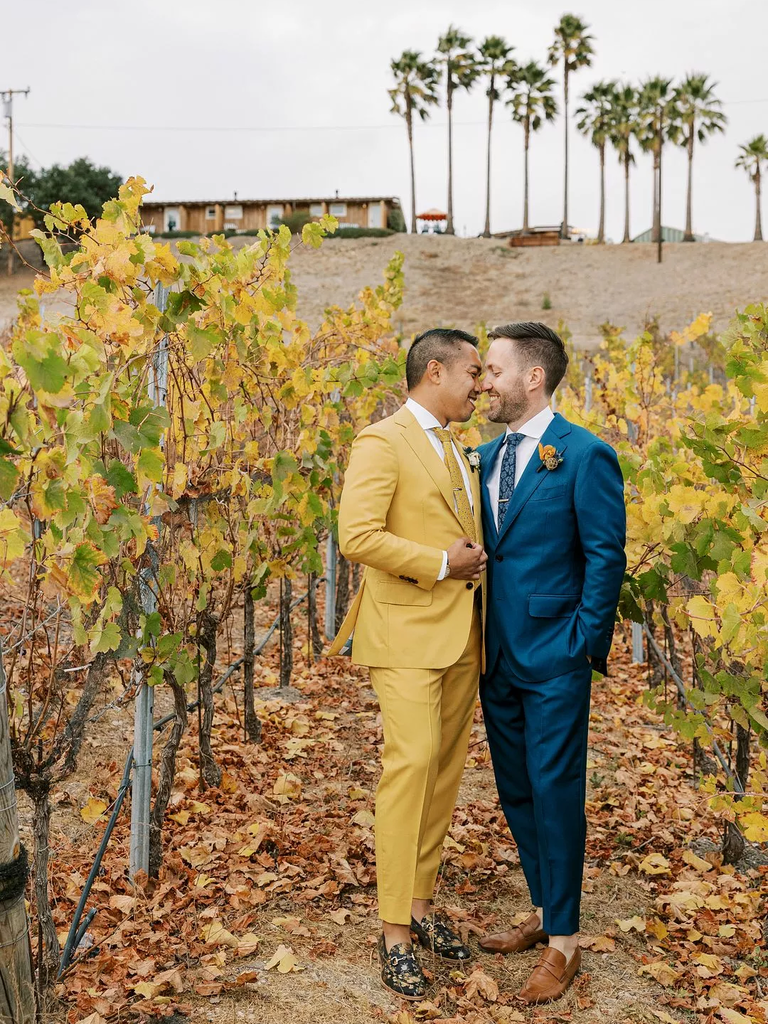A pair of grooms lean in for a kiss among the yellows and green of an autumnal winery.