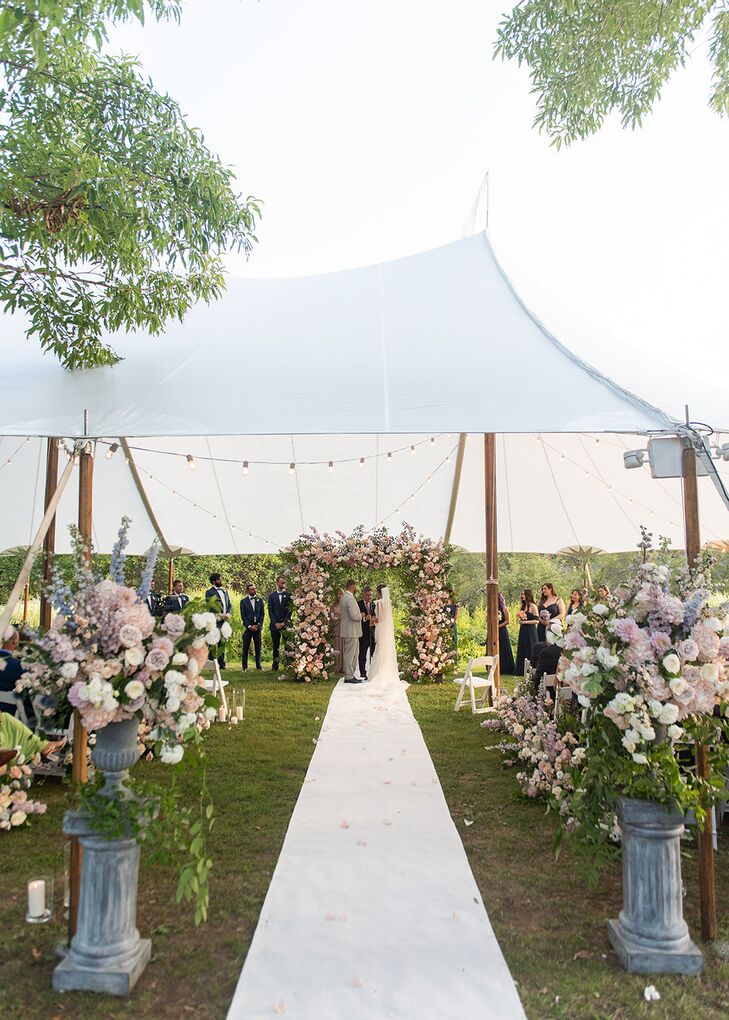 The bride and groom say their vows under a lush pastel chuppah at the end of a white aisle lined with large floral arrangements with light blooms under a tent and string lights at a farm venue.