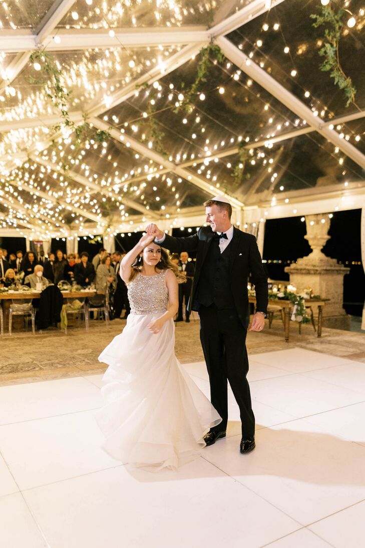 Groom in Black Tuxedo, Kippah Twirls Bride in Tulle A-Line Gown on Dance Floor Under Tent, String Lights