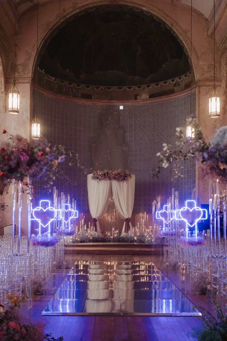A whimsical church-like ceremony space at Hotel Peter and Paul in New Orleans, LA, set up to resemble Baz Luhrmann's "Romeo and Juliet" starring Leonardo DiCaprio and Claire Danes with a mirrored aisle, tall and painted floral arrangements, neon heart crucifixes, drapery and many candles at the altar.