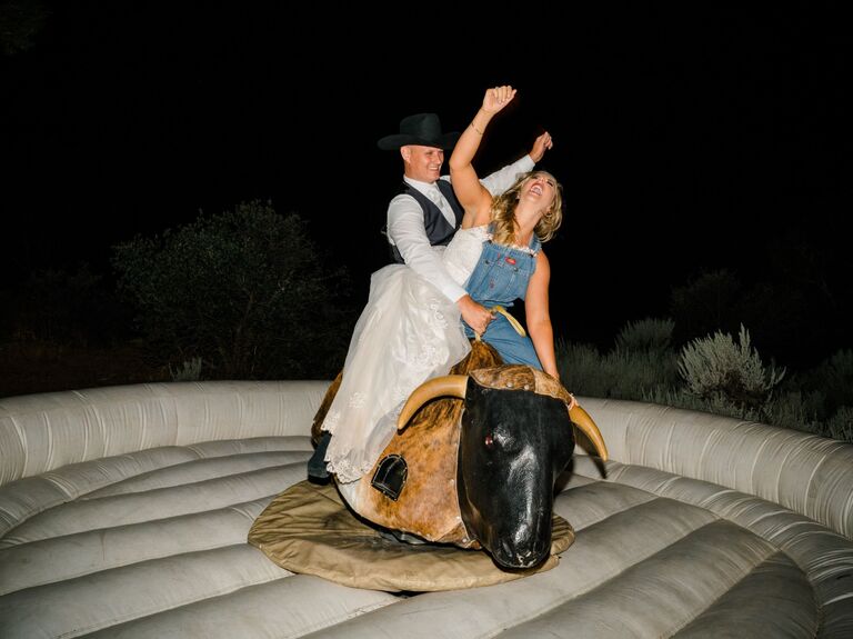 Couple rides a mechanical bull while laughing and waving their arms in the air. 