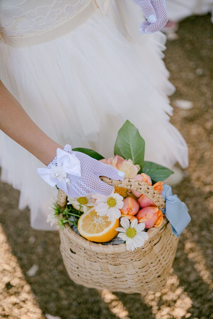 A flower girl with a delicate mesh gloves and a bow holding a wicker basket filled with daisies, buds, greenery and a half of a lemon for an outdoor garden wedding in California.