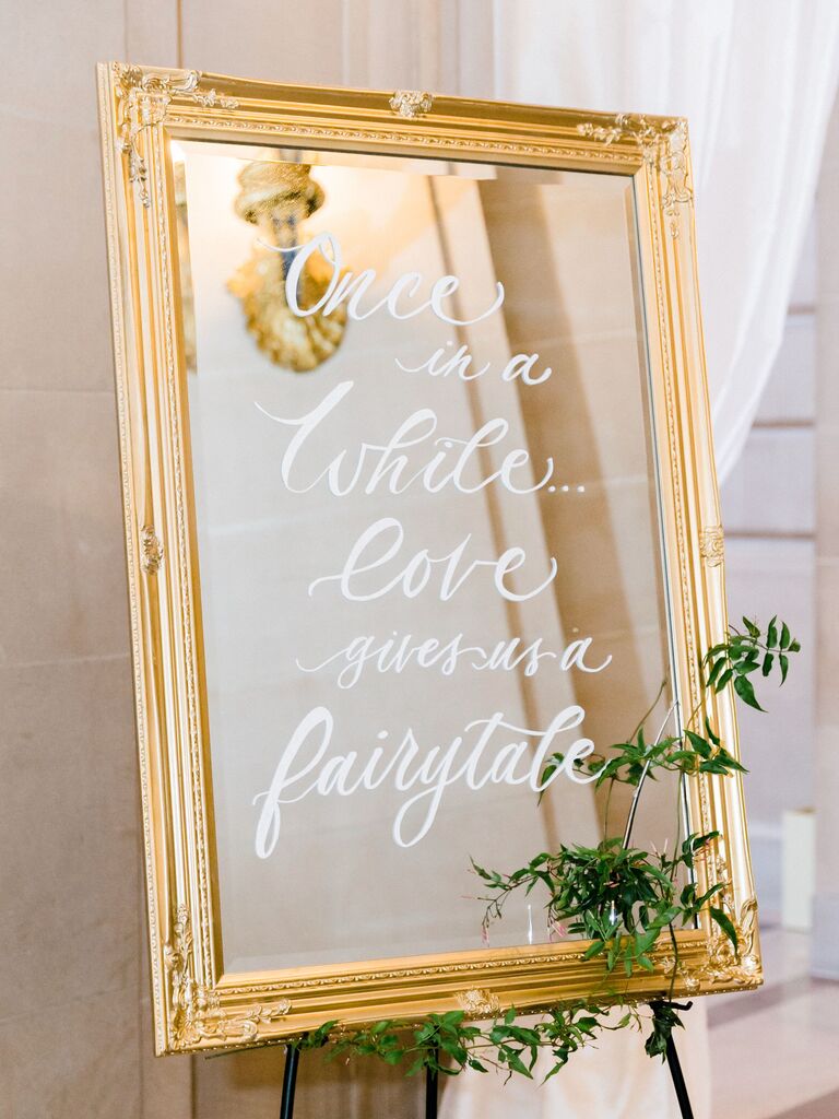 mirror wedding sign in gold frame and quote written in white calligraphy: "once in a while, love gives us a fairytale"