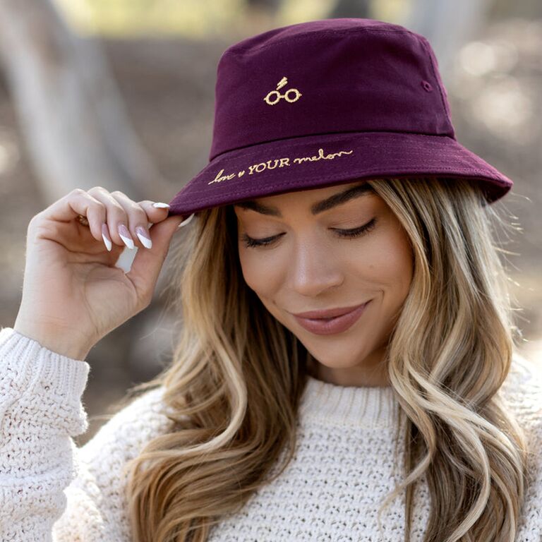 A burgundy bucket hat with a gold embroidered Harry Potter glasses and scar design and cursive brand name from Love Your Melon