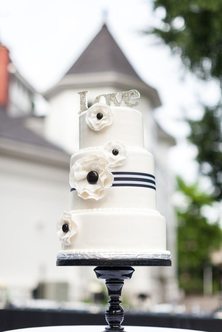 The main ivory wedding cake stood three tiers tall and had black-and-white sugar flowers decorating the surface. The middle layer of the cake had a black-and-white-striped ribbon wrapped around the middle. The cake was topped with a silver sparkly  "Love" sign, and the entire dessert was positioned on a simple black cake stand.