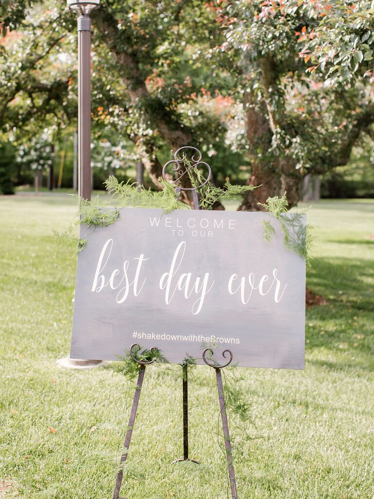 Wedding welcome sign on metal stand in the grass with couple's wedding hashtag