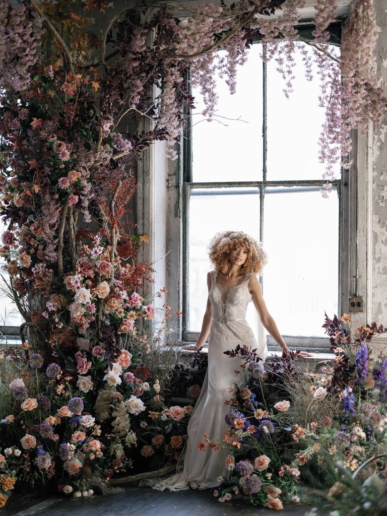 2023 wedding flower trend bride stands against a large window underneath a canopy of purple and blush flowers