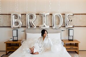 Bride in Satin Robe Getting Ready on Wedding Morning With Balloons Spelling "BRIDE"