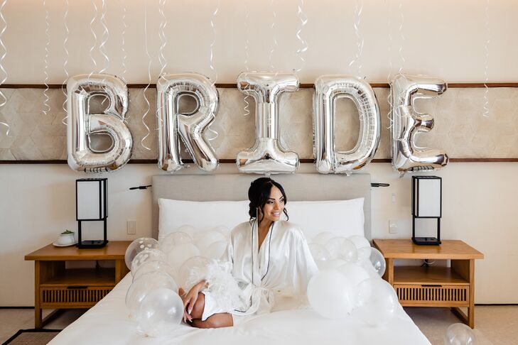 A bride gets ready in her hotel room before her destination wedding in Los Cabos, Mexico, with a satin robe, a beautiful updo hairstyle and a large balloon sign before her that reads "BRIDE."
