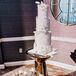 A four-tier white wedding cake with edible floral details on a sleek and shiny gold cake stand with a fresh arrangement of white-and-blush arrangement at a luxurious wedding.