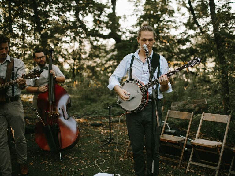Musician plays the banjo in an outdoor wooded area. 