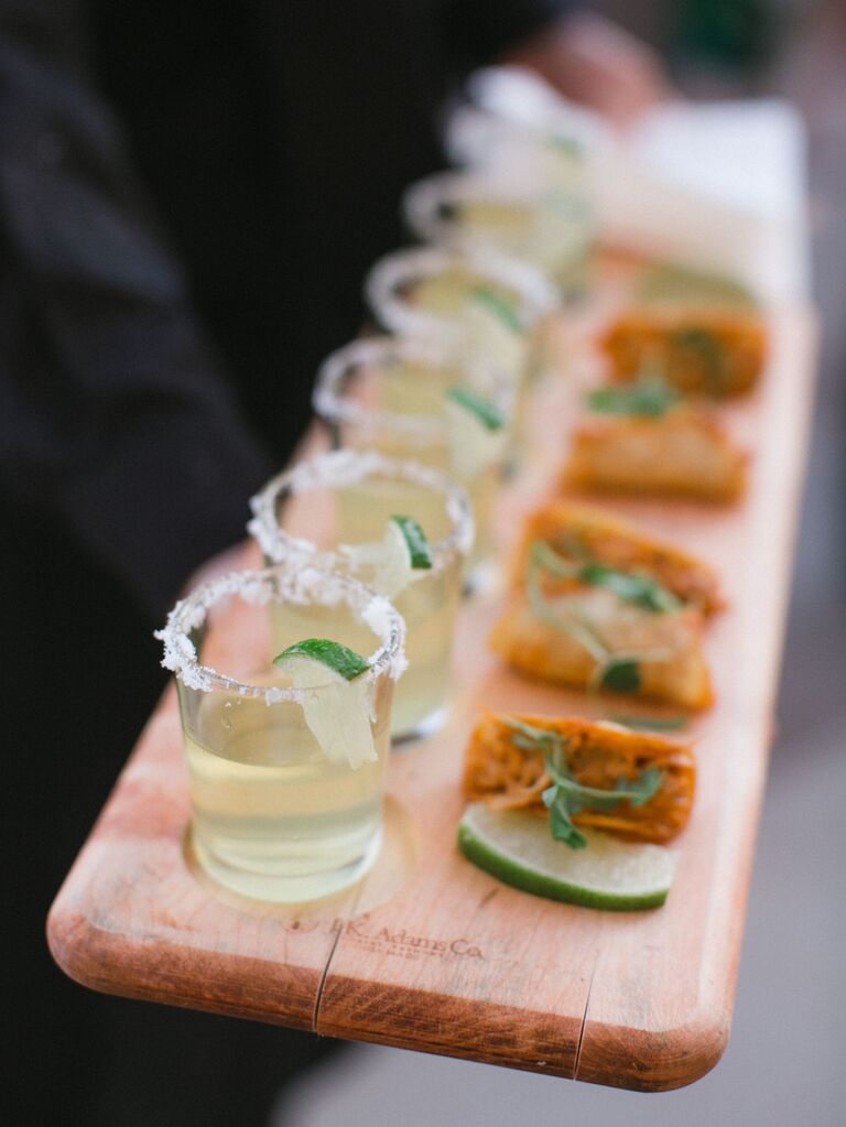 Board displays mini glasses of salt-rimmed margaritas and crispy tacos. 
