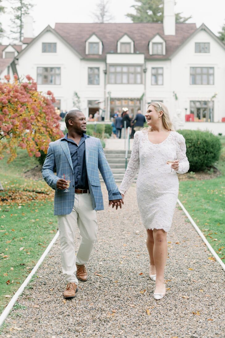 A bride in a shortened version of her mother's own wedding dress with lace and beading, a simple down hairstyle and a pearl headband with her groom in a blue, plaid suit jacket and a blue dress shirt and light pants walking hand-in-hand outside of their autumn rehearsal welcome dinner in the Berkshires.