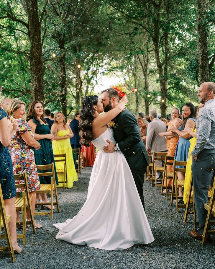 Bride in Sleek A-LIne Dress and Groom in Gray Kissing Halfway Up Aisle Under Tree Canopy