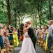 Bride in Sleek A-LIne Dress and Groom in Gray Kissing Halfway Up Aisle Under Tree Canopy