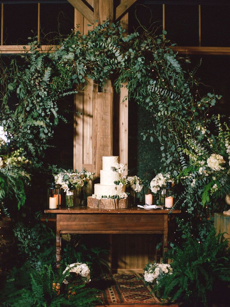 Three-tier rustic wedding cake on tree stump stand under greenery arch