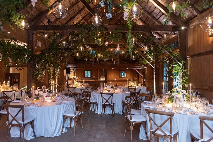 A barn reception space on a farm with hanging greenery, lavender flowers and large bulbs over round tables with crisp white linens, low centerpieces and romantic candles at an elegant, rustic-chic wedding.