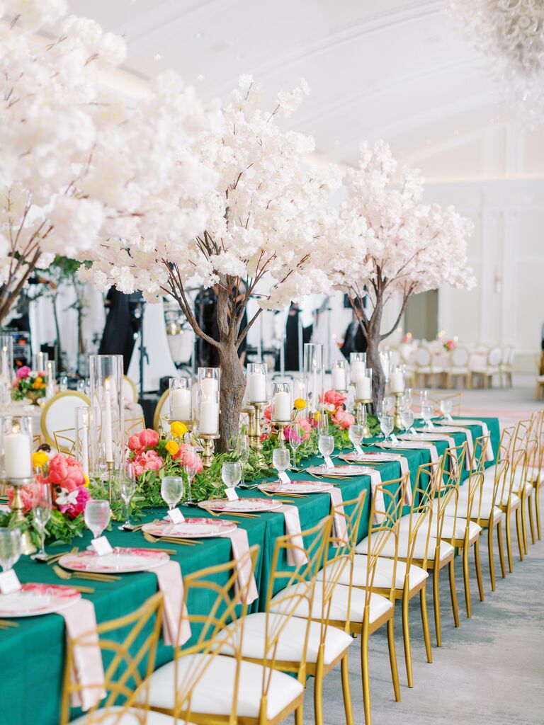 long wedding reception table with emerald green tablecloth, low centerpieces of pink and orange flowers with tall white cherry blossom trees down the middle of the table