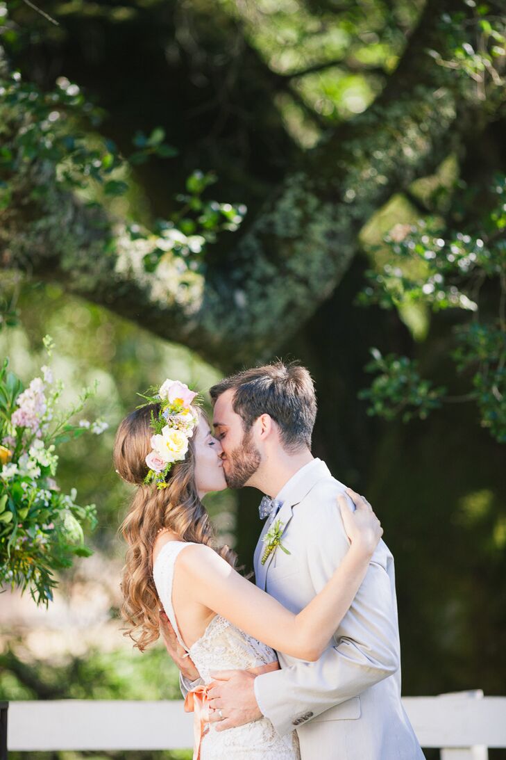 Doug wore a tan suit on the wedding day, with a white button-up collared dress shirt and blue bow tie accented with white. Holly kissed him while she wore her pastel flower crown, created with peonies and roses and accented with fern. She wore a custom-made lace wedding dress with sleeves, and had a peach-colored sash wrapped around her waist.