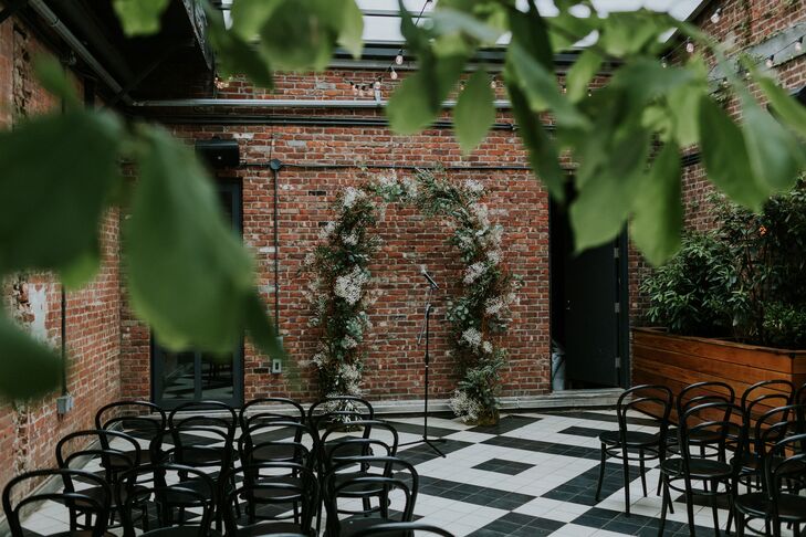 An industrial ceremony space with a wedding arch featuring white flowers and greenery, a black-and-white floor, exposed bring and string lights in New York City.
