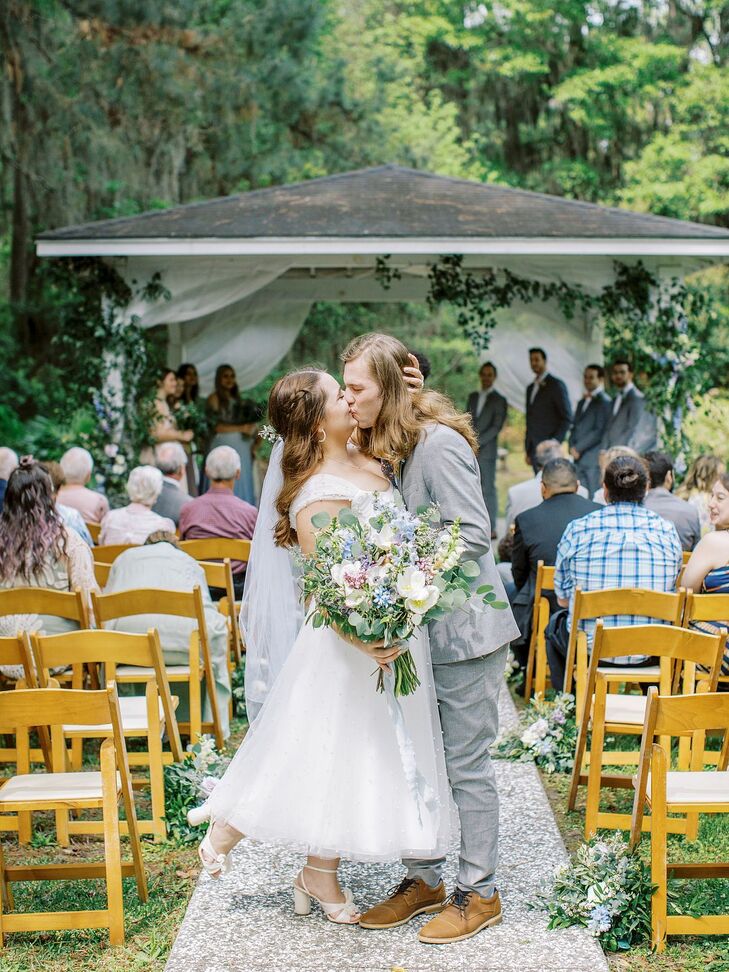 A couple share a kiss after exchanging wedding vows under a gazebo in a beautiful garden setting at their vintage and floral-inspired wedding.