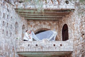 Groom in White and Black Suit Jacket Embraces Bride in Fitted Gown and Veil Blowing in the Wind on Castle Balcony