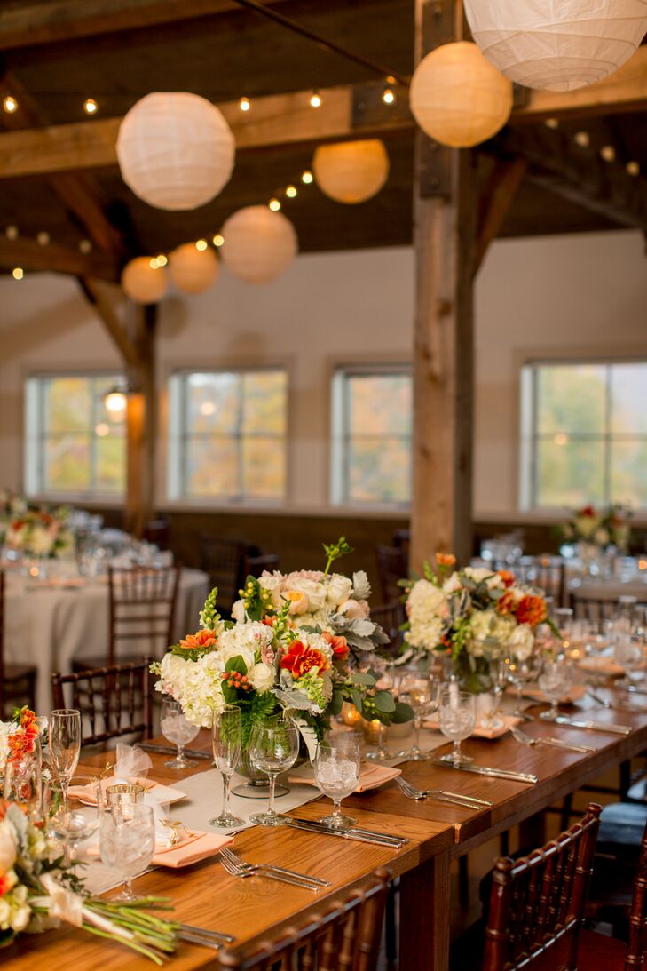 Wooden farm tables with neutral table runners and brown chiavari chairs added to the rustic ambience inside barn reception at the Mountain Top Inn & Resort in Chittenden, Vermont. Low centerpieces of ivory and orange flowers continued the fall theme, and paper lanterns and string lights overhead brought out a romantic ambience.