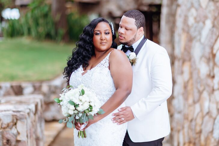 Groom in White and Black Tux Jacket and Boutonniere Holds Bride in Fitted Wedding Dress With Lace and Deep V-Neckline, White Bouquet, Outside Castle