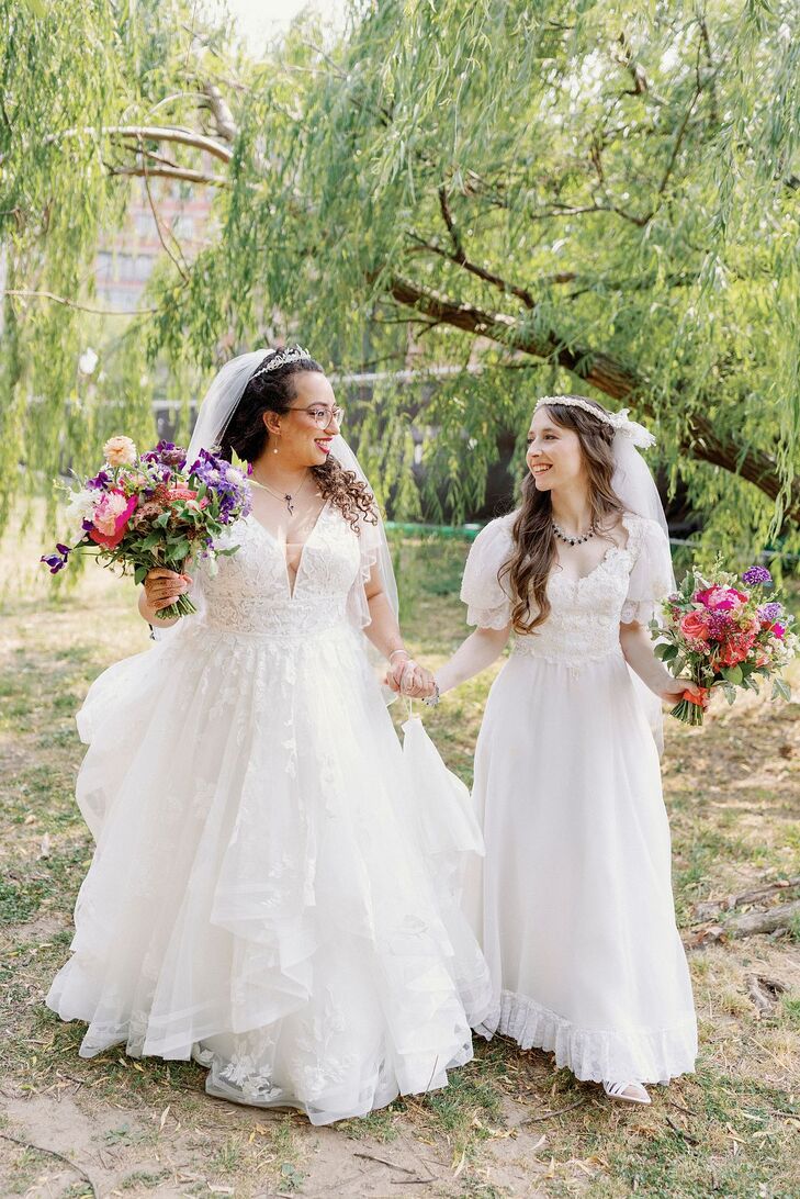 A bride in an a-line gown with a plunging neckline and floral detailing with a headpiece and a purple-and-coral bouquet holding hands with her bride in a vintage gown that her mother wore on her wedding day, as well as a retro headpiece and veil and a pink bouquet.