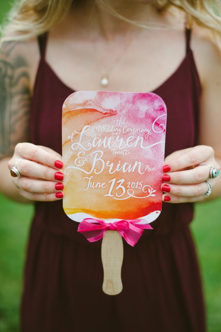 Wedding programs depicted on decorative fans had pink and orange watercolor motifs seen throughout the day's paper goods. The props had pink bows tied at the bottom near the wooden handle, which guests gripped to fan themselves throughout the summer ceremony.