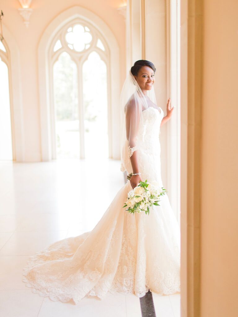 bride wearing lace fit-and-flare dress with train stands in chapel corridor