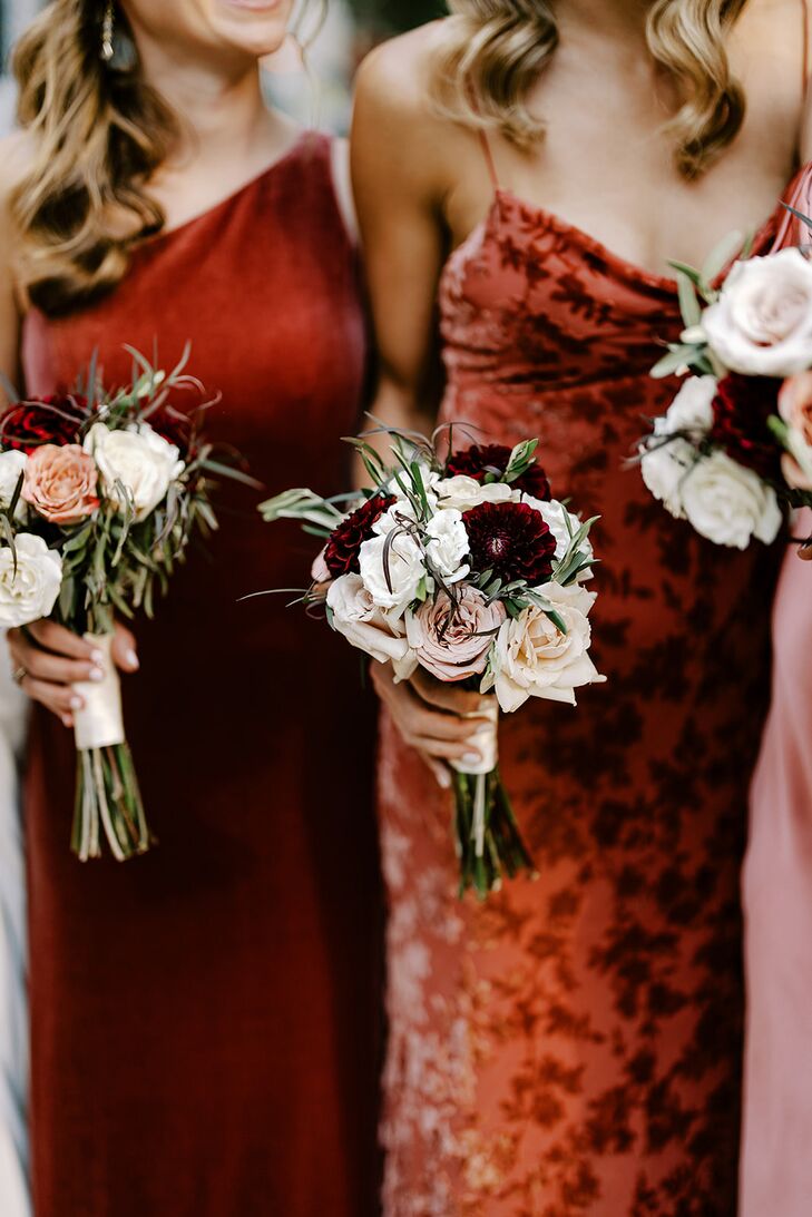 Small nosegay bouquets with roses and other flowers in deep red, white and blush colors accented with greenery held by bridesmaids in different red, pink and champagne-colored dresses for a classic and elegant wedding.