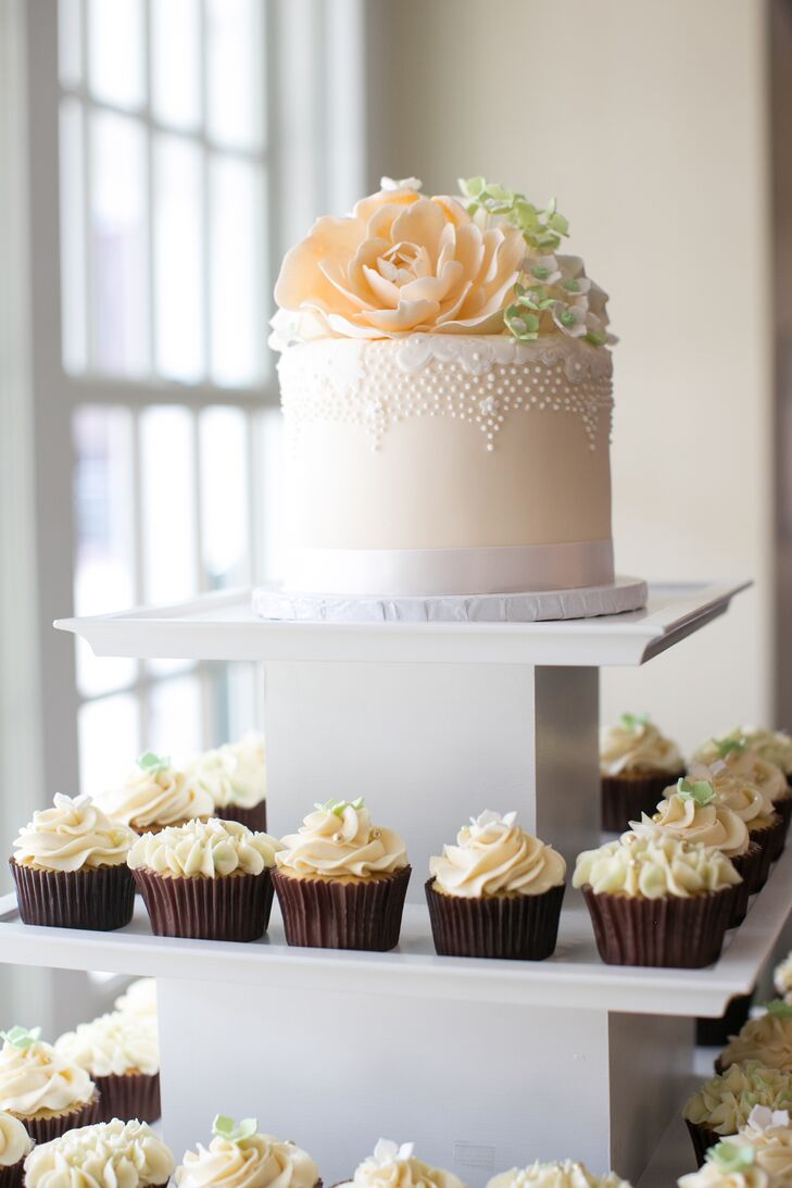 The simple ivory wedding cake was topped with peach and green cake flowers, which stood on top of an assortment of buttercream frosted cupcakes.