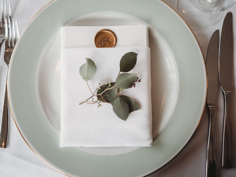 wedding place setting with sage green dinner plate, white menu card folded into napkin and sprig of fresh eucalyptus