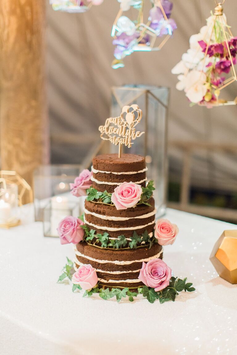 three tier chocolate naked wedding cake decorated with ivy vines, pink roses and gold cake topper that says our adventure awaits