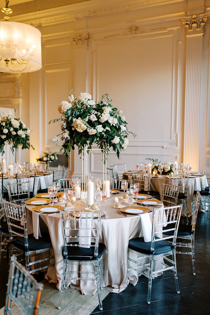 Round reception tables with silk gray linens, candles and classic arrangements of white flowers and greenery suspended from gold structures surrounded by ghost Chiavari chairs in an elegant ballroom.