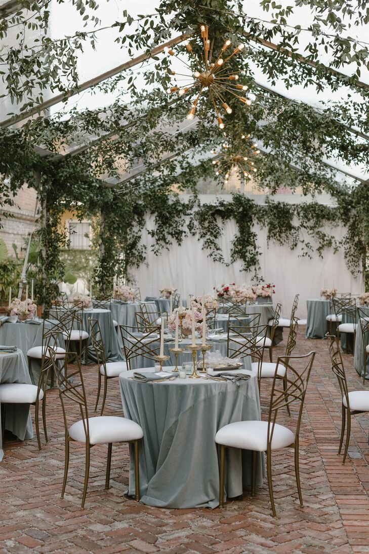 A tented reception space outside Hotel Peter & Paul with hanging greenery, modern chandeliers and small cafe tables with blue linens, gold candles and low centerpieces with pink and blush flowers.