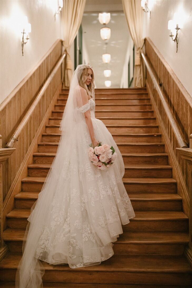 A bride with a classic and elegant gown--off-the-shoulder, A-line dress with lace detailing poses on the stairs of her venue with a crystal crown, a long veil and a pink bouquet.