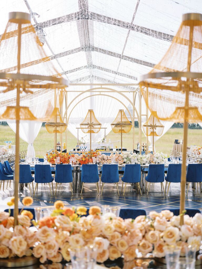 cleartop wedding reception tent with chandeliers, orange centerpieces and velvet royal blue chairs along tables