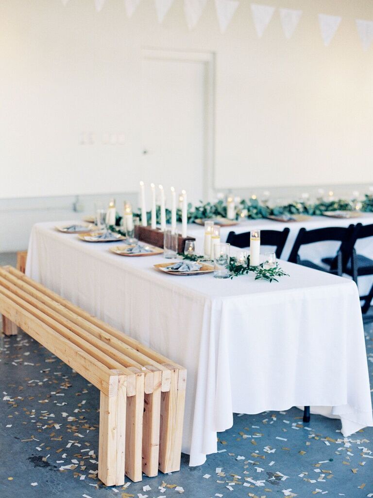 minimalist wedding table decor with white tablecloth, white taper candles in wooden holder, greenery table runner and long wooden bench