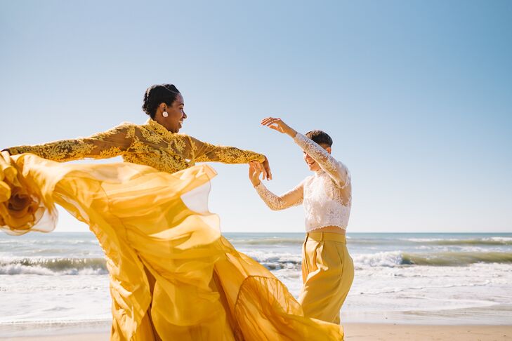 A bride in a yellow, two-piece outfit with a lace bodice and a flowing skirt and her partner in a white lace bodice and yellow slacks frolicking and dancing during their first look on the beach.