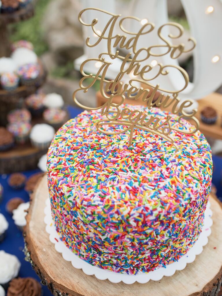 wedding cake with rainbow sprinkles and gold cake topper that says "and so the adventure begins"