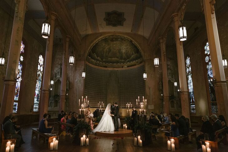 A romantic and dimly lit ceremony space at Hotel Peter & Paul with church detailing, stained-glass windows, low and tall candles, greenery and pews in the round surrounding the bride and groom on a raised platform.