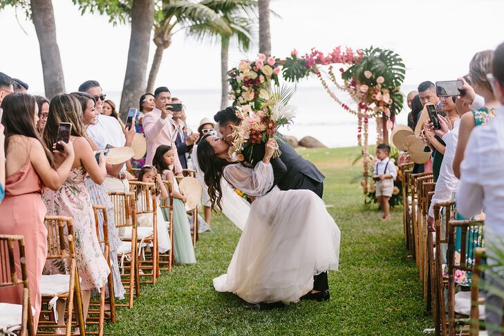 A groom in a black tuxedo dips his new bride, wearing a fitted gown with illusion details, into a kiss during their ceremony recessional at their bohemian wedding with tropical details and bright colors in Hawaii.