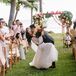 A groom in a black tuxedo dips his new bride, wearing a fitted gown with illusion details, into a kiss during their ceremony recessional at their bohemian wedding with tropical details and bright colors in Hawaii.