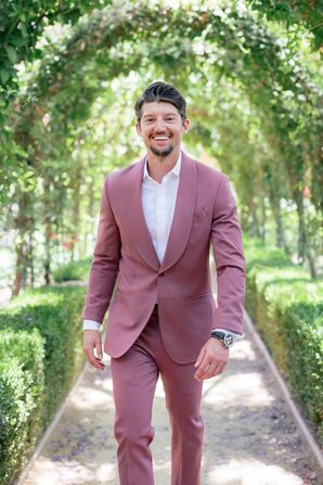 Groom in Dusty Rose Pink Suit and White Accents Under Greenery Arches